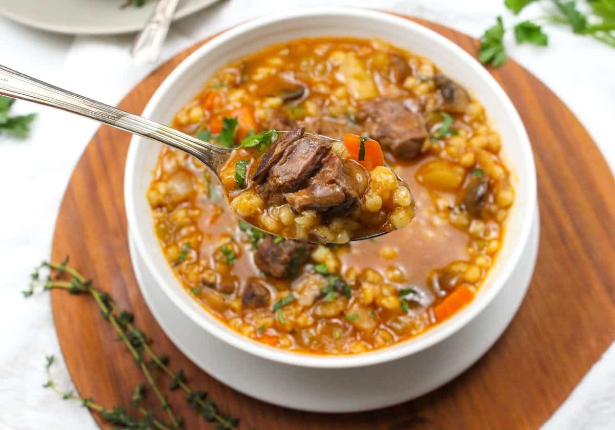 A spoonful of beef and barley soup with a bowl in the background.