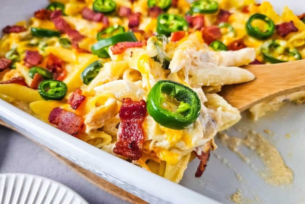 A serving spoon scoops into a baked casserole with penne pasta, cheese, jalapeños, and bacon pieces.