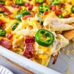 A serving spoon scoops into a baked casserole with penne pasta, cheese, jalapeños, and bacon pieces.