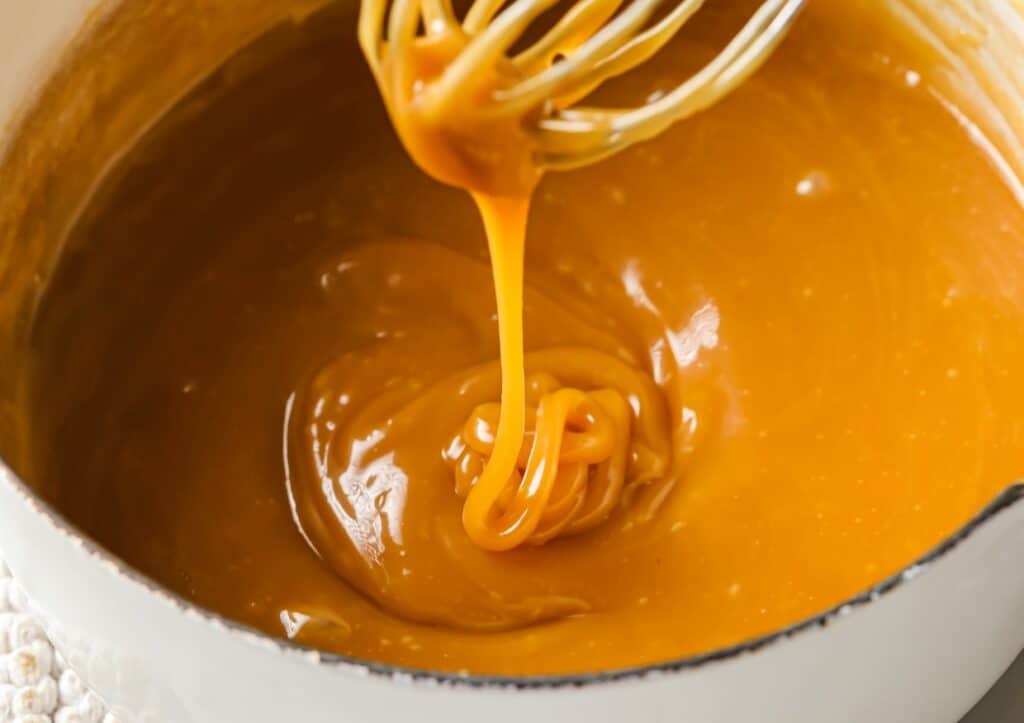 Whisk lifting a thick, golden caramel sauce from a white pot.