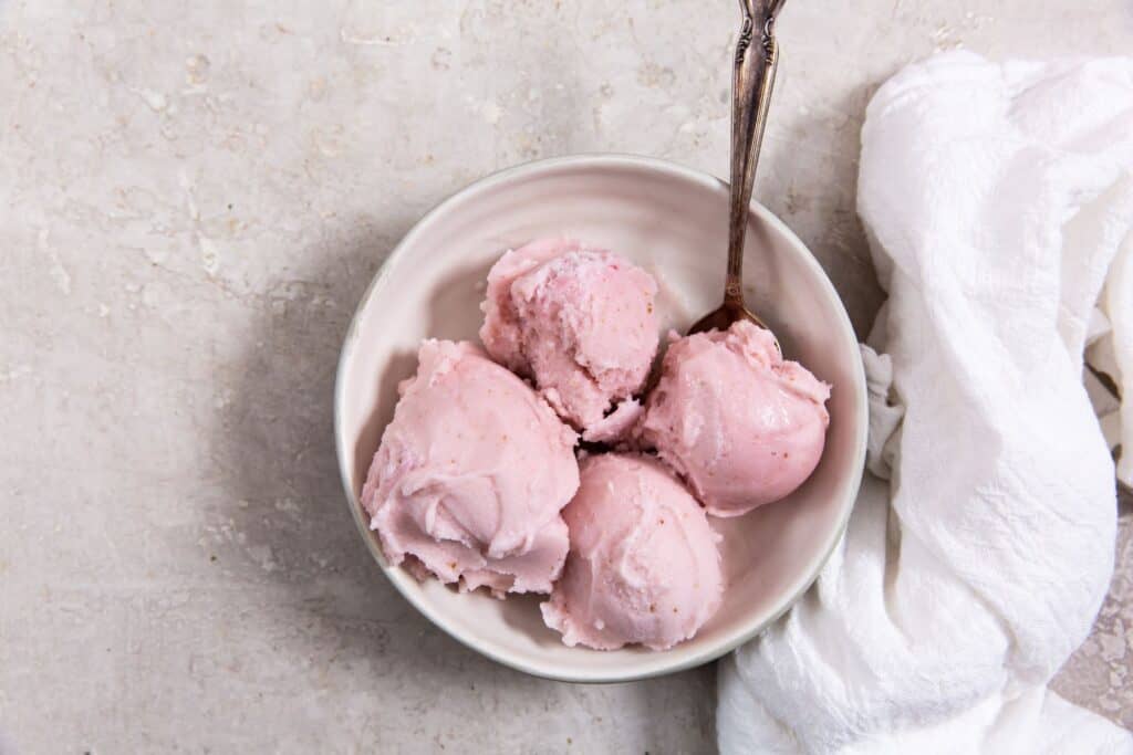 Bowl of strawberry ice cream with three scoops and a spoon, on a concrete surface with a white cloth nearby.