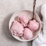 Bowl of strawberry ice cream with three scoops and a spoon, on a concrete surface with a white cloth nearby.