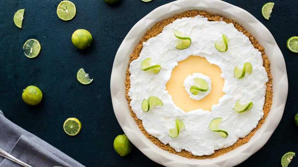 A key lime pie with whipped cream and lime zest garnish, surrounded by fresh limes on a dark surface.