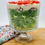 A layered salad in a clear glass trifle bowl on a wooden board, with peas, lettuce, and red tomatoes, next to a floral patterned cloth.