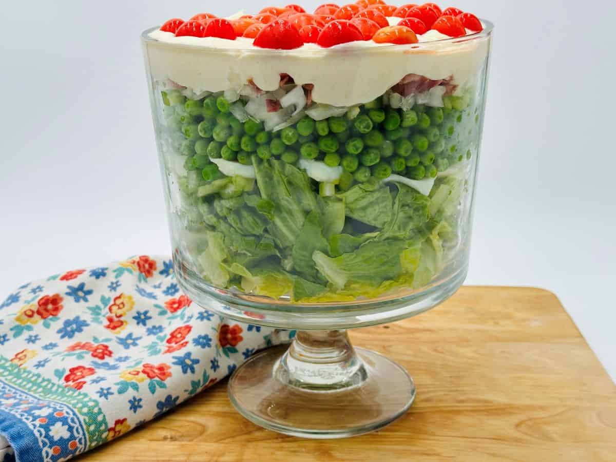 A layered salad in a clear glass trifle bowl on a wooden board, with peas, lettuce, and red tomatoes, next to a floral patterned cloth.