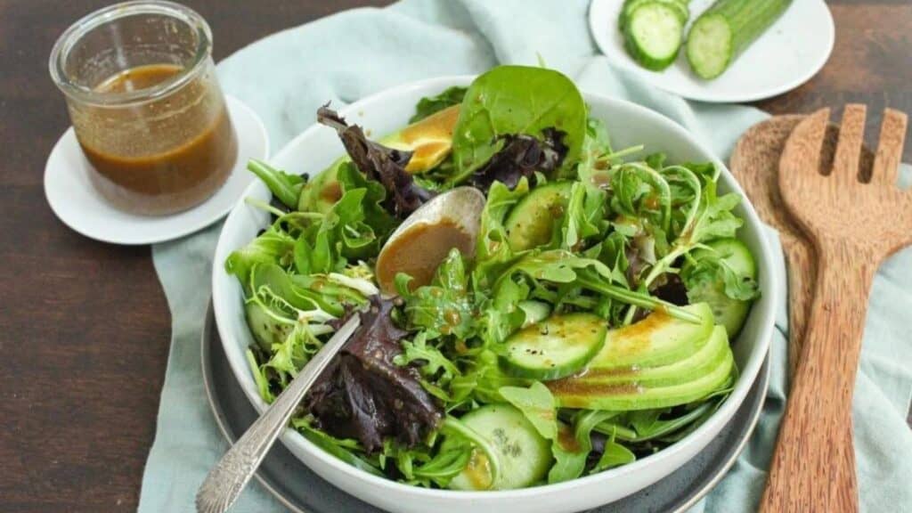 A bowl of fresh green salad with arugula, spinach, cucumber slices, and avocado, served with a side of dressing.