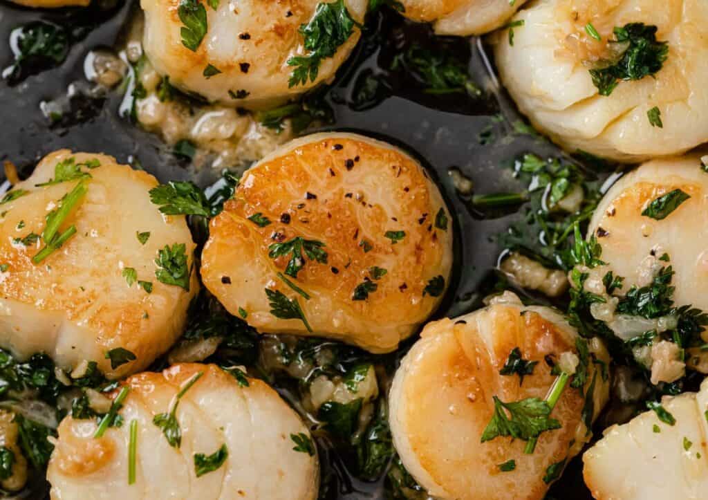 Pan-seared scallops with golden crusts, garnished with chopped parsley, in a black skillet.