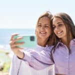 Two smiling women taking a selfie with a smartphone by the seaside on a sunny day.