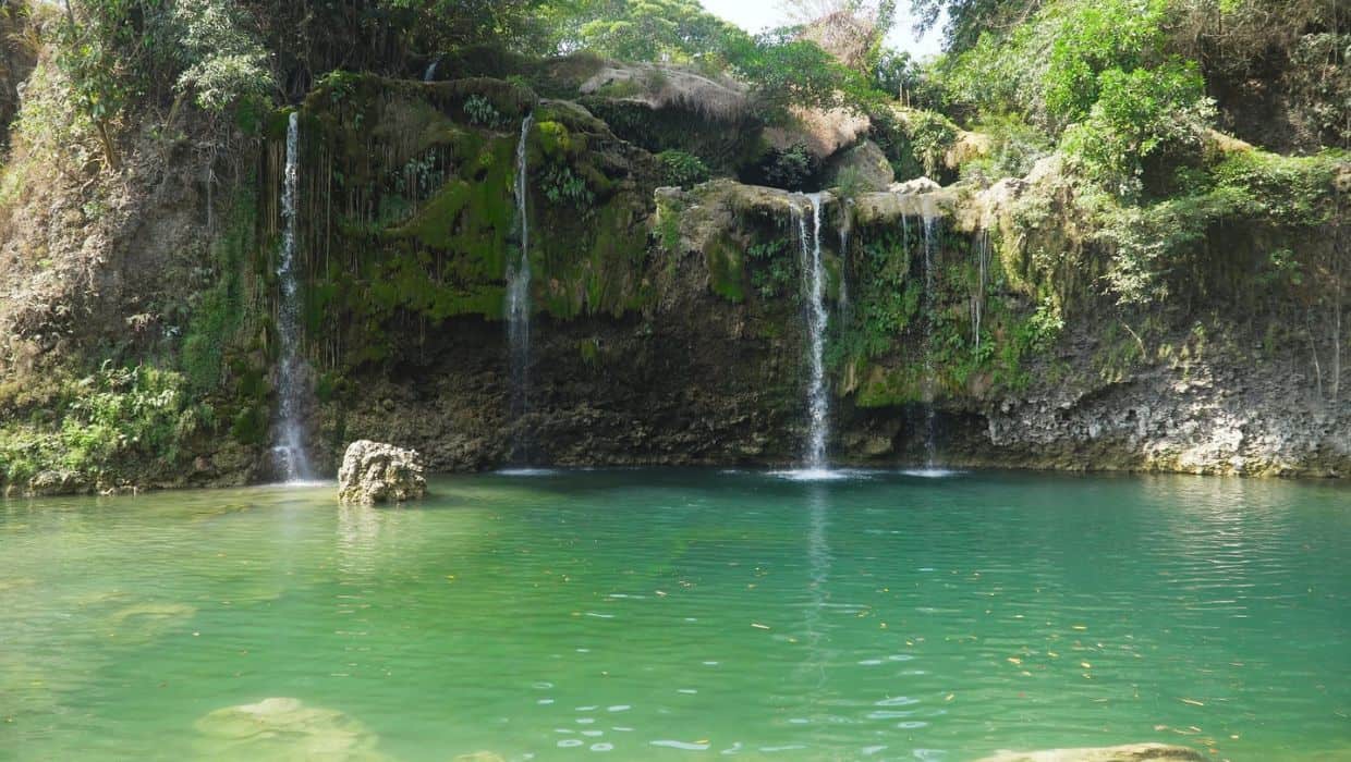A serene waterfall, one of the must-visit places in the Philippines, flows into a tranquil emerald green pool surrounded by lush greenery.