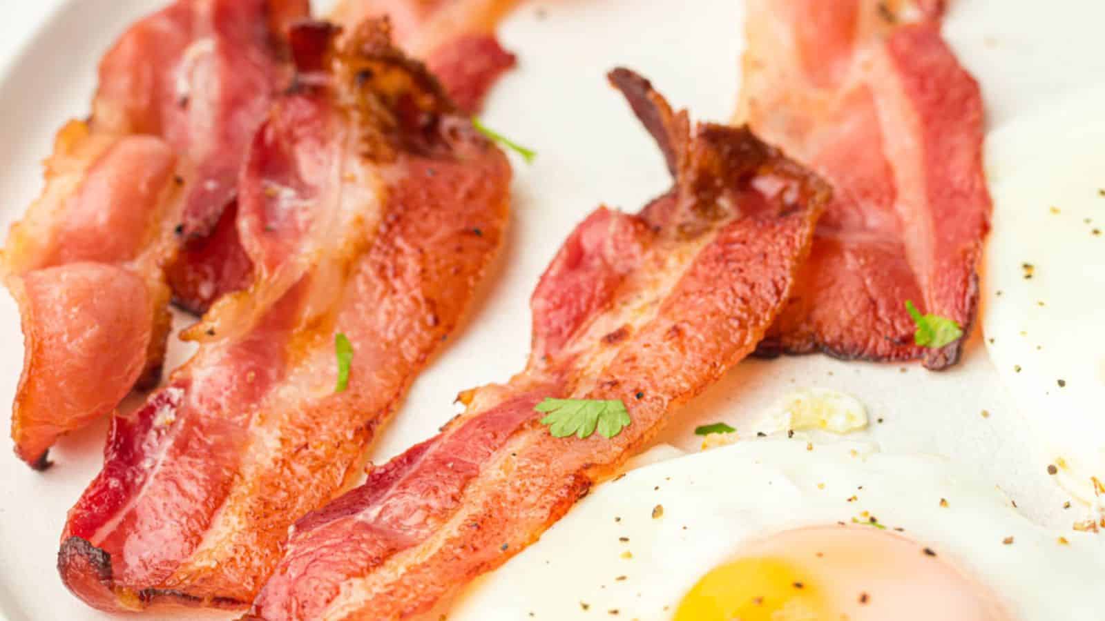 Close-up view of cooked bacon strips and fried eggs on a white plate, garnished with parsley.