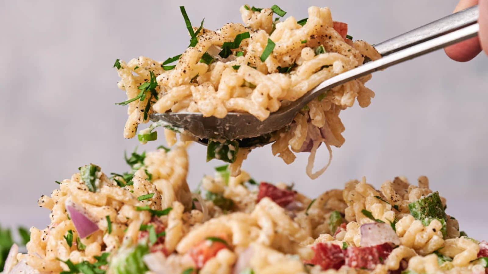A spoon lifting a serving of creamy macaroni salad with diced red onion and fresh herbs, with pepper sprinkled on top.
