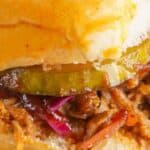 Close-up of a pulled pork sandwich with pickles and red cabbage on a bun, held in hand, with chips in the background.