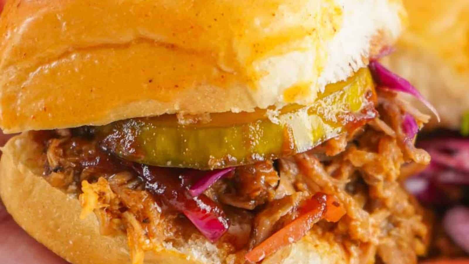 Close-up of a pulled pork sandwich with pickles and red cabbage on a bun, held in hand, with chips in the background.