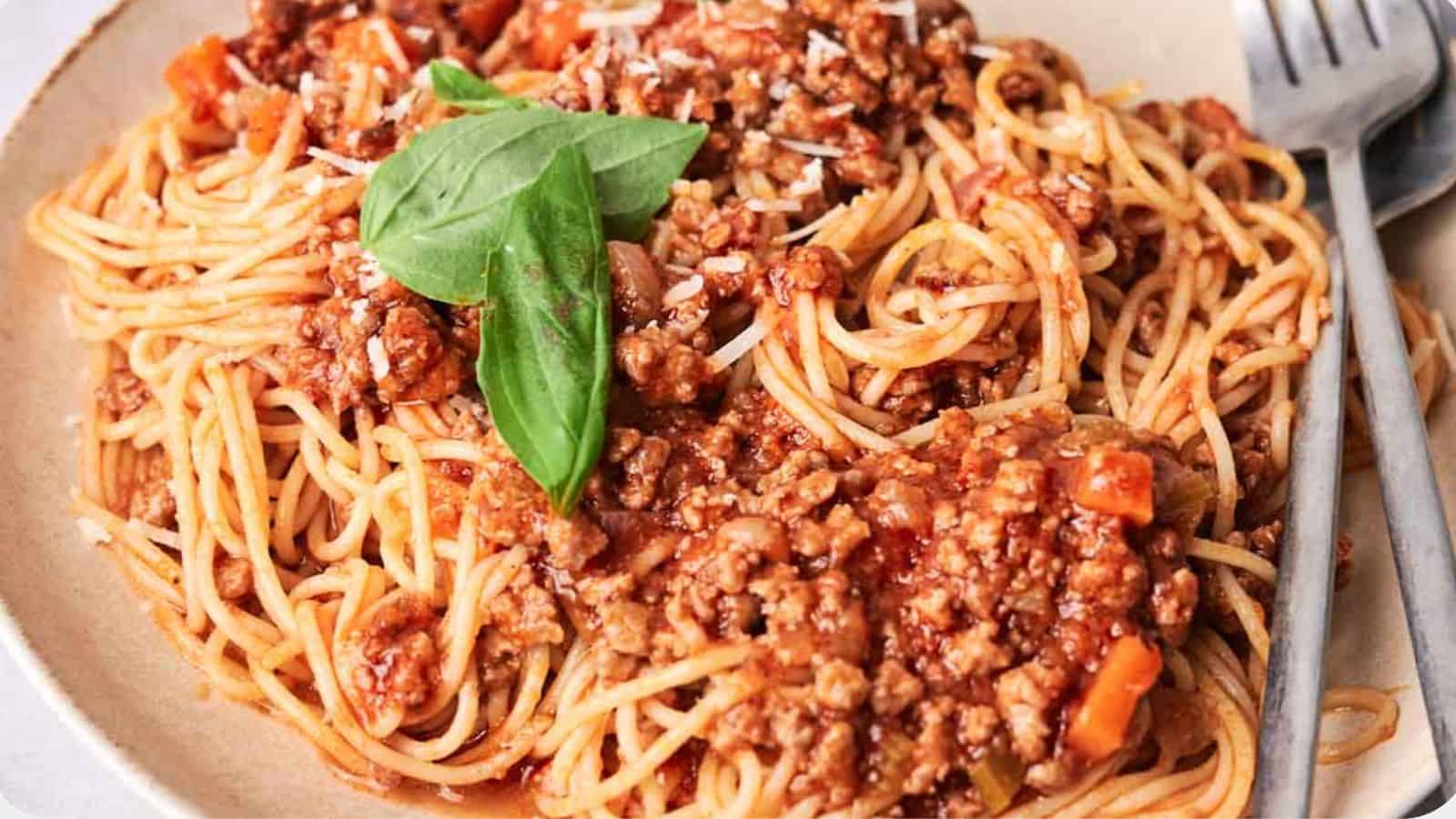 A plate of spaghetti topped with meat sauce and a fresh basil leaf, accompanied by a fork on the side.