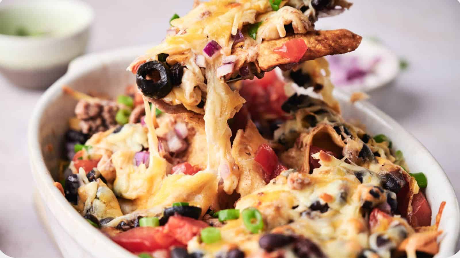 A bowl of taco casserole topped with melted cheese, black olives, tomatoes, and green onions, with a creamy dip in the background.