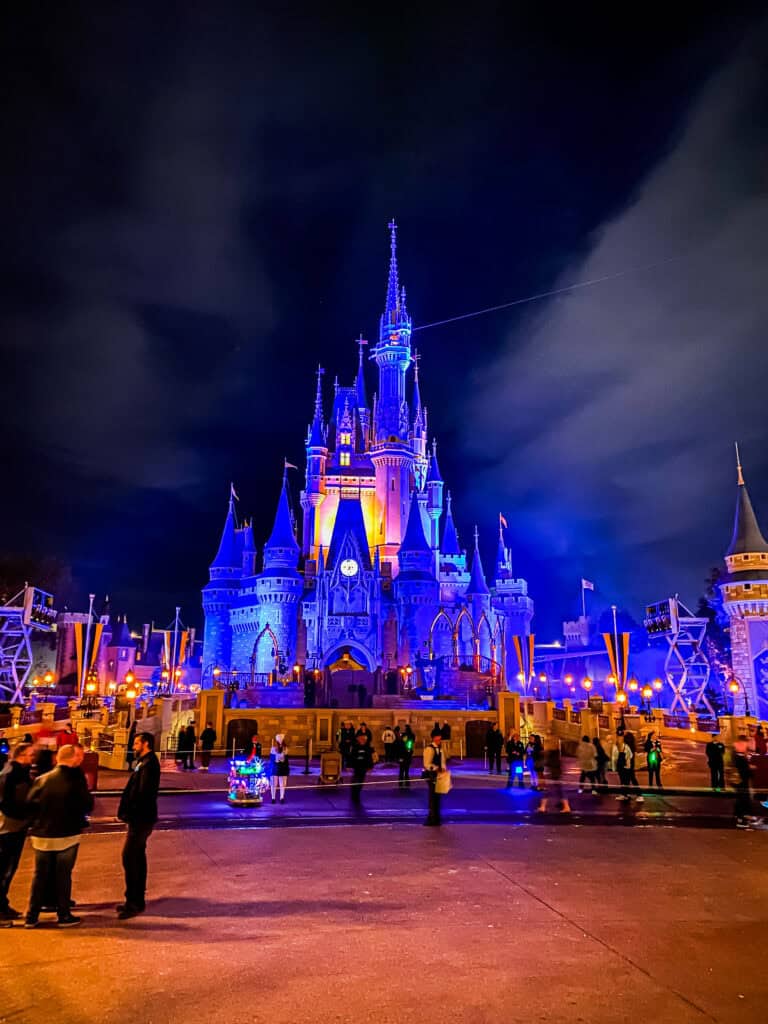 Night view of cinderella's castle illuminated in blue and purple lights at a theme park, with visitors visible in the foreground.