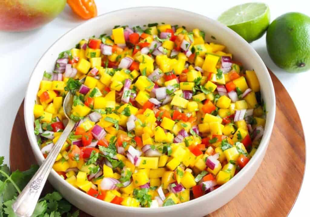 A bowl of fresh mango salsa with chopped onions, peppers, and cilantro, accompanied by a lime and bell pepper on a wooden board.