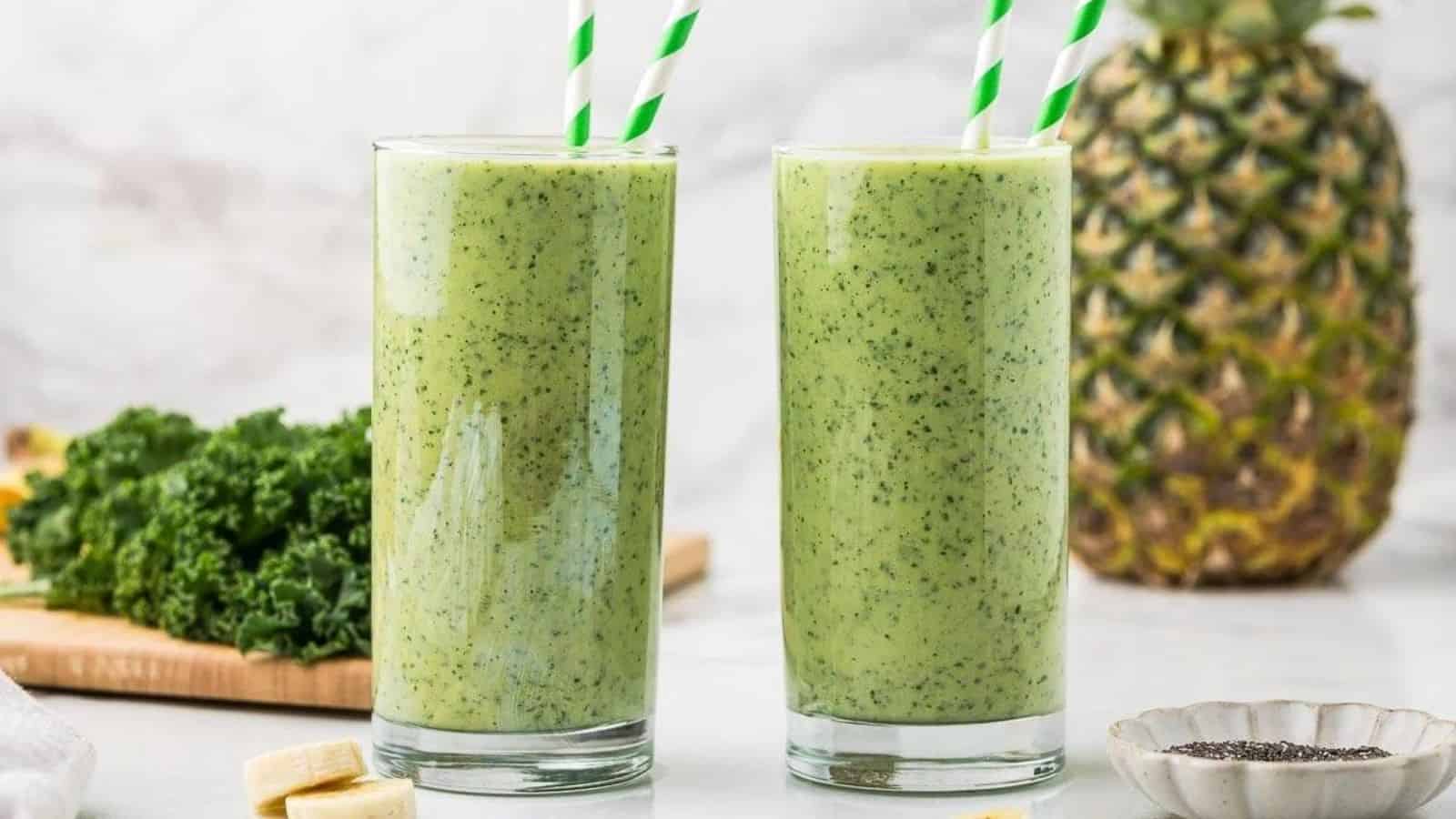 Two green smoothies in tall glasses with striped straws with a pineapple behind it and a wooden cutting board with kale on top.