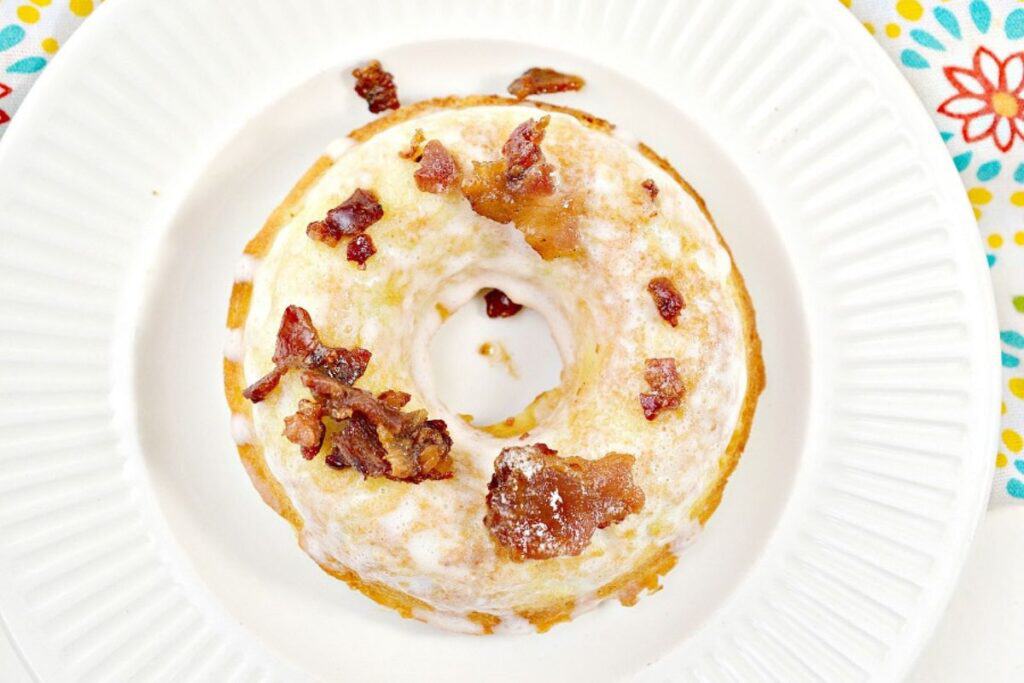 A glazed donut topped with bacon pieces on a white plate with a floral pattern.