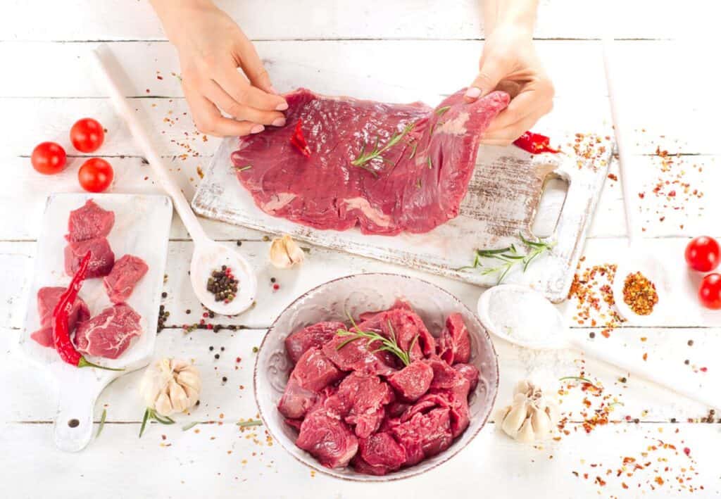 A person preparing meat with herbs on a kitchen counter, surrounded by ingredients and spices.