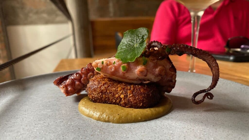 Gourmet dish featuring a grilled octopus tentacle served on a crispy croquette with a dollop of sauce, presented on a ceramic plate with a diner in the background.