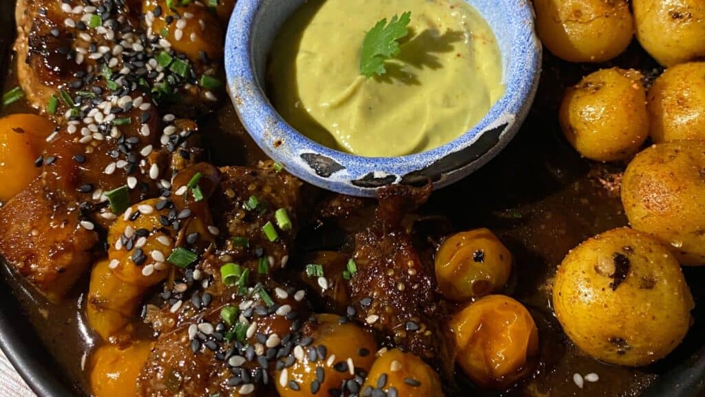 A dish of glazed meat with sesame seeds, accompanied by round roasted potatoes and a bowl of green sauce.