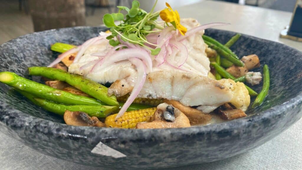 Grilled fish fillet served with asparagus, corn, mushrooms, and garnished with onion and edible flowers in a stone bowl.