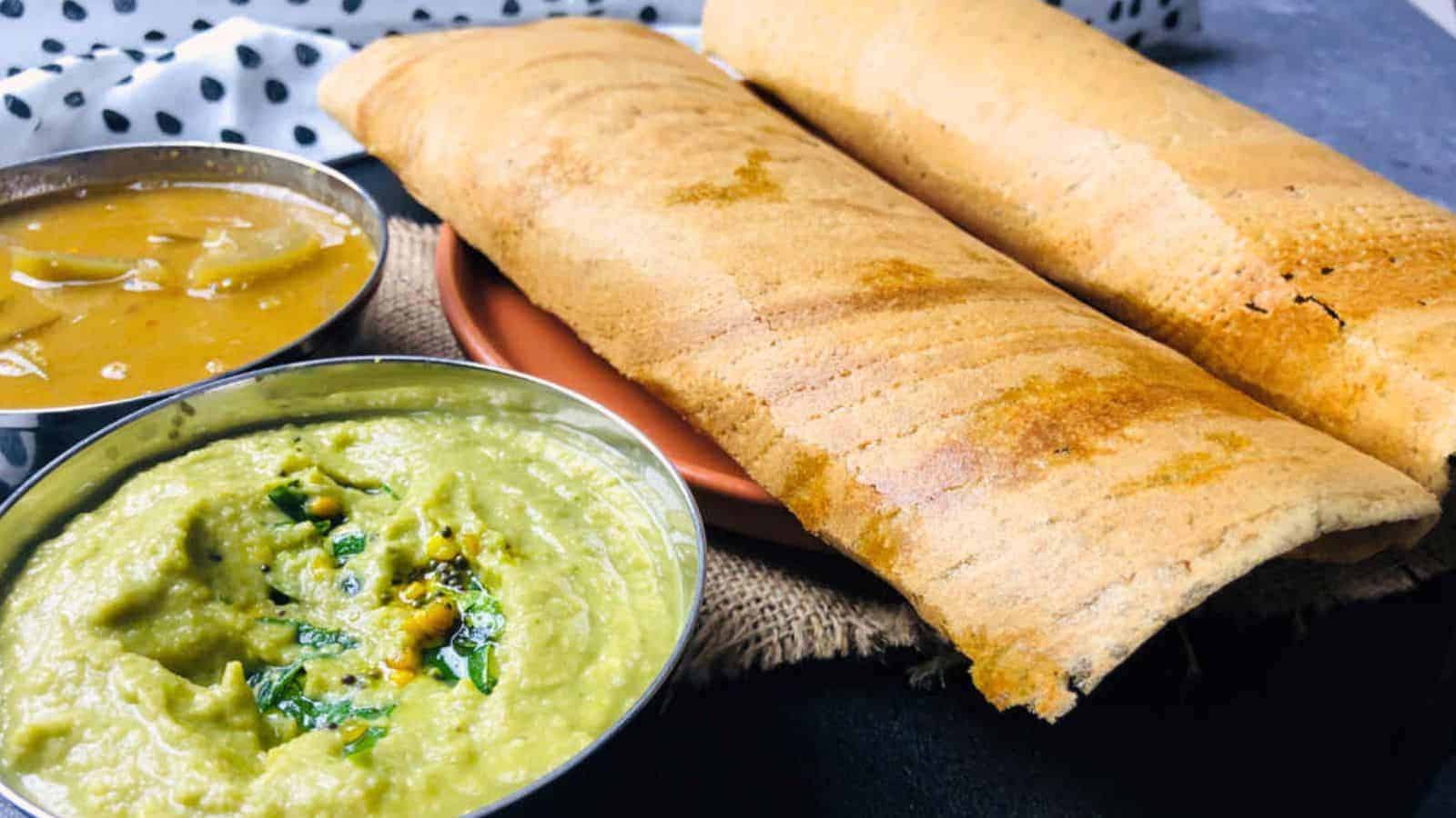 Two dosas served with coconut chutney and sambar in small bowls, placed on a rustic fabric surface.