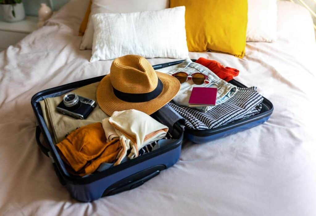 Open suitcase on a bed with neatly packed items including clothes, a hat, sunglasses, and a camera, suggesting preparations for a trip.