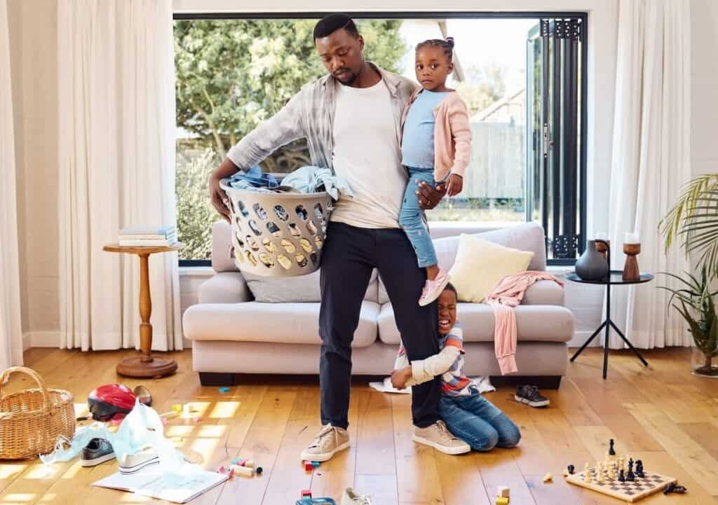 A man holding a laundry basket balances a toddler on his hip while another child playfully clings to his leg in a messy living room.