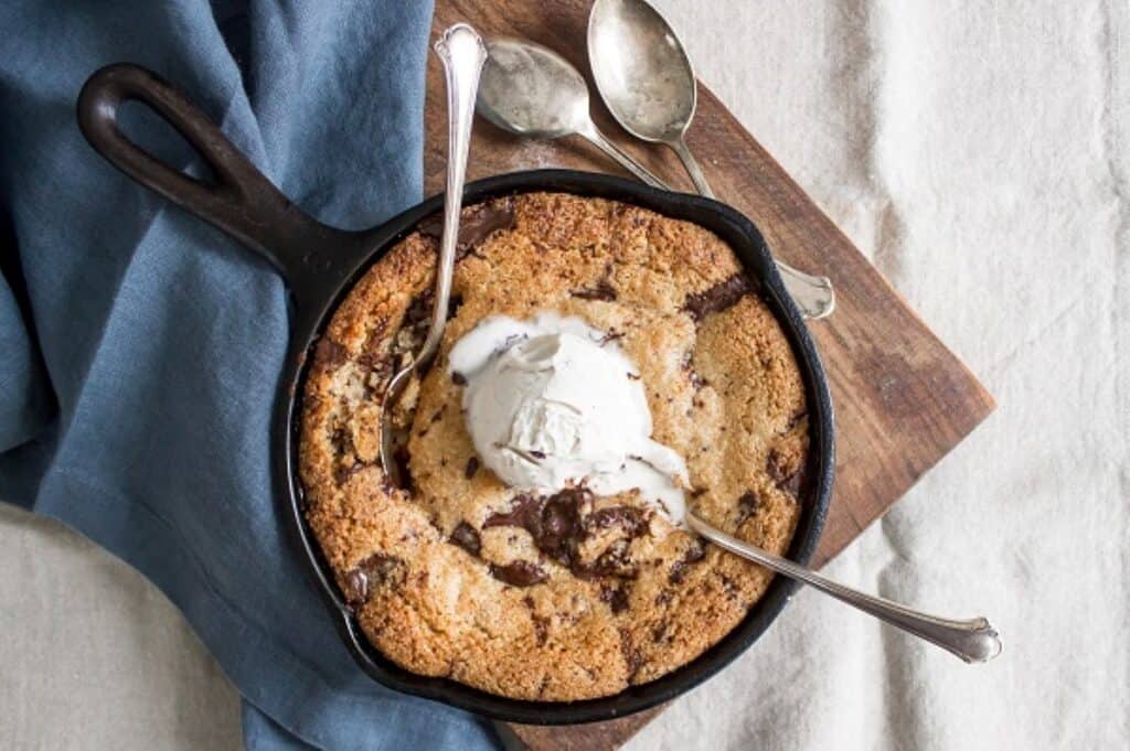 A freshly baked skillet cookie topped with a scoop of vanilla ice cream, accompanied by spoons ready for serving.