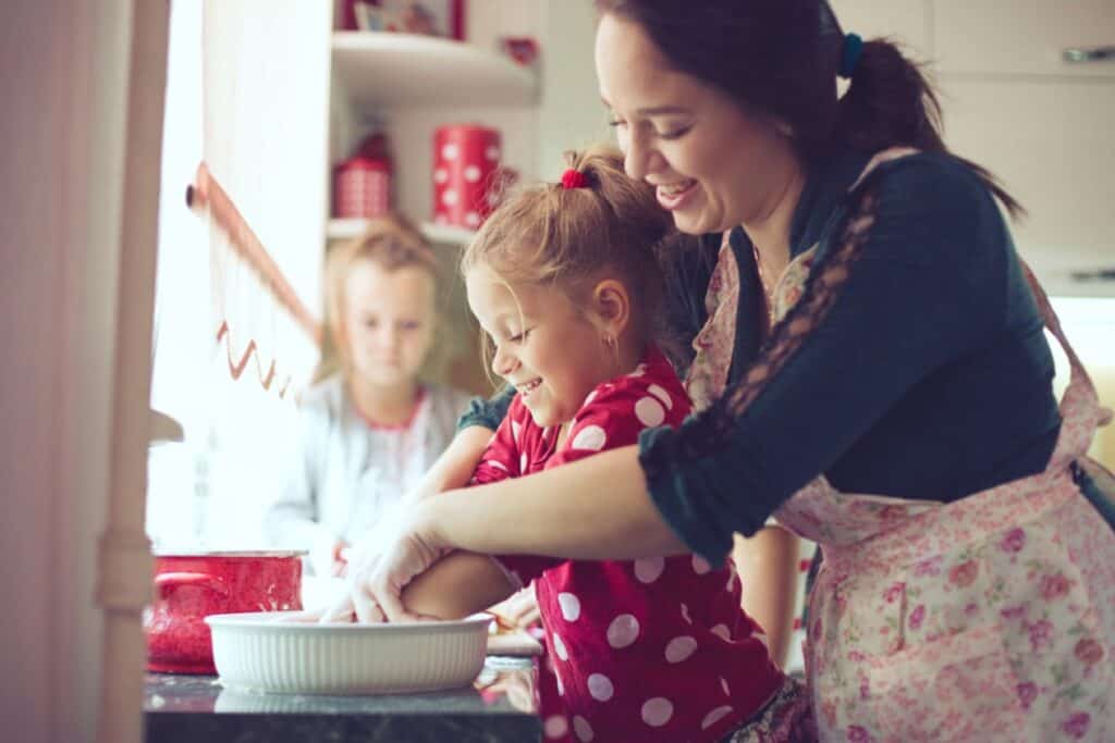 A smiling woman teaching two young girls to bake in a home kitchen.
