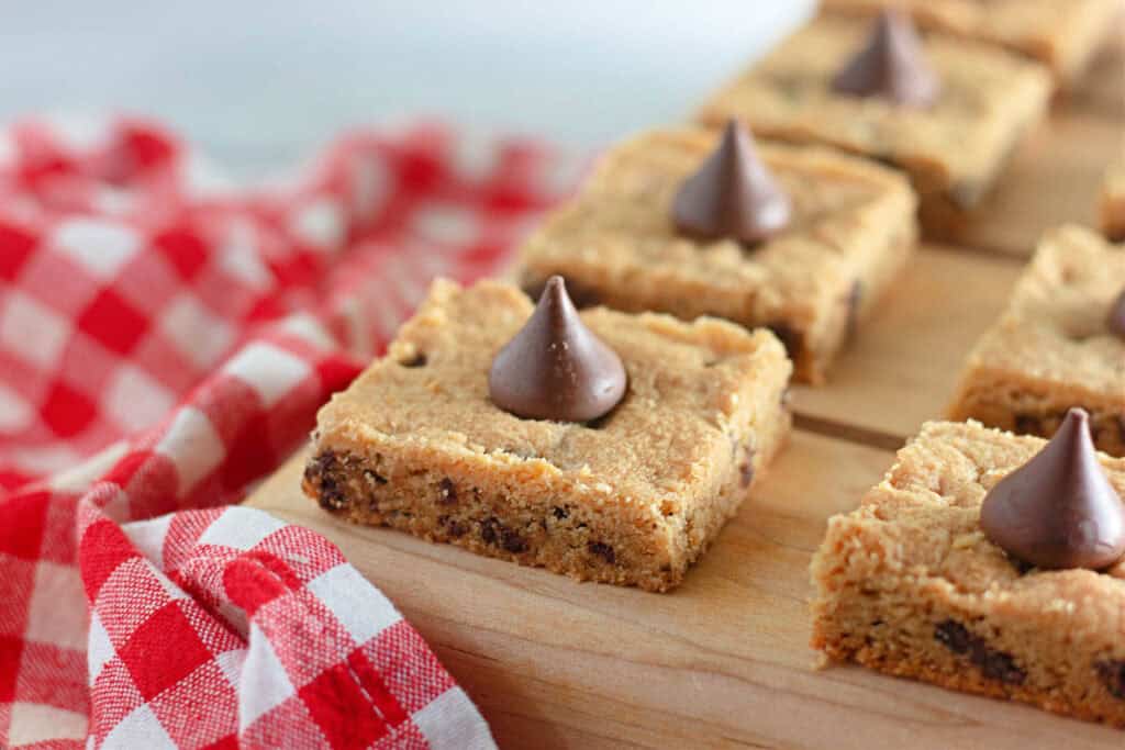 Freshly baked cookie bars topped with chocolate kisses, displayed on a wooden board beside a red and white checkered napkin.