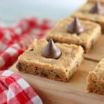 Freshly baked cookie bars topped with chocolate kisses, displayed on a wooden board beside a red and white checkered napkin.