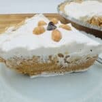 A slice of peanut butter pie topped with whipped cream and peanuts, on a white plate with the whole pie in the background.