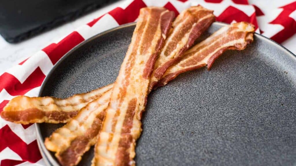 Crispy bacon strips on a round plate with a red and white napkin in the background.