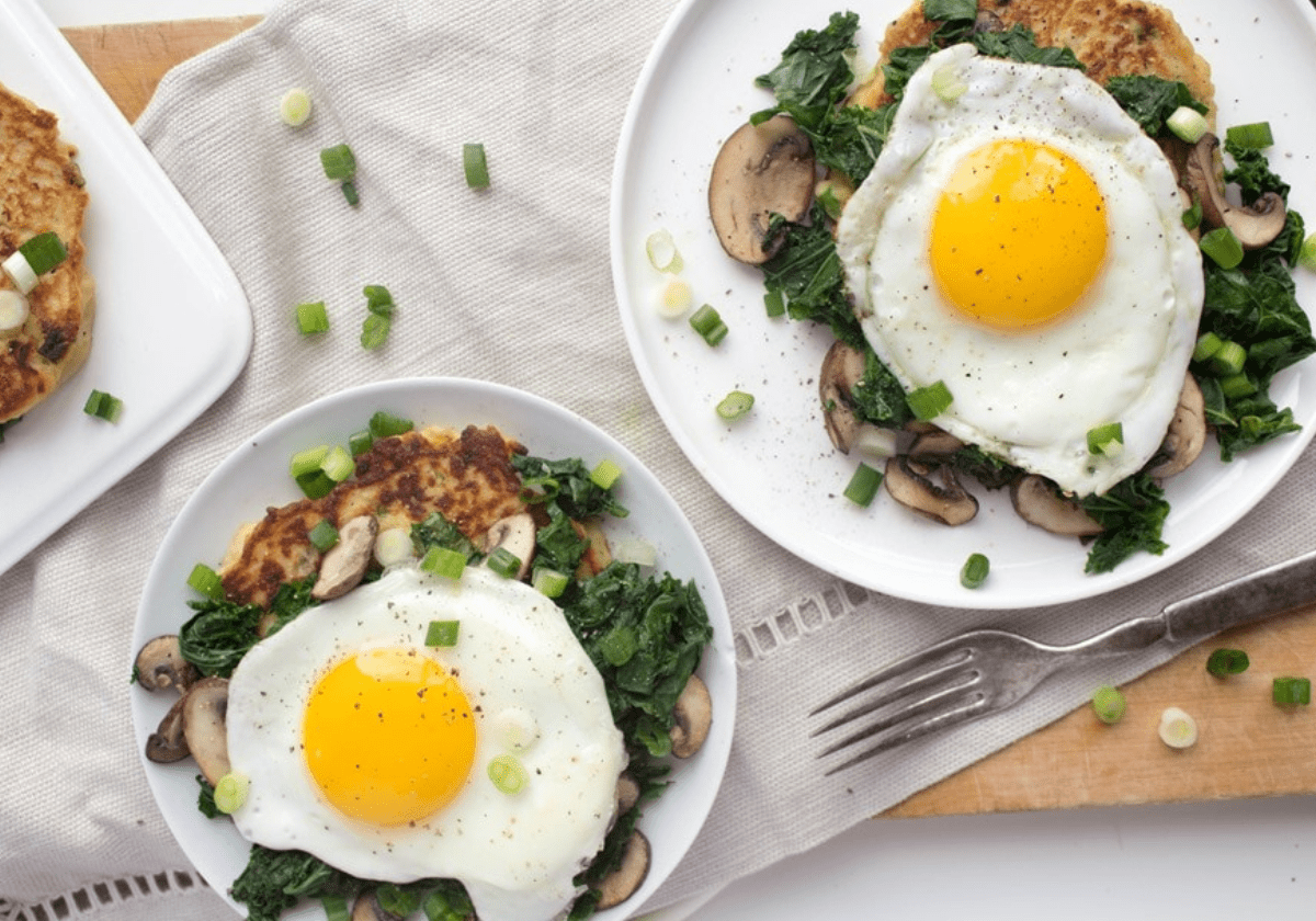 Two mashed potato pancakes on white plates topped with fried eggs, sauteed mushrooms, spinach, and fresh herbs.