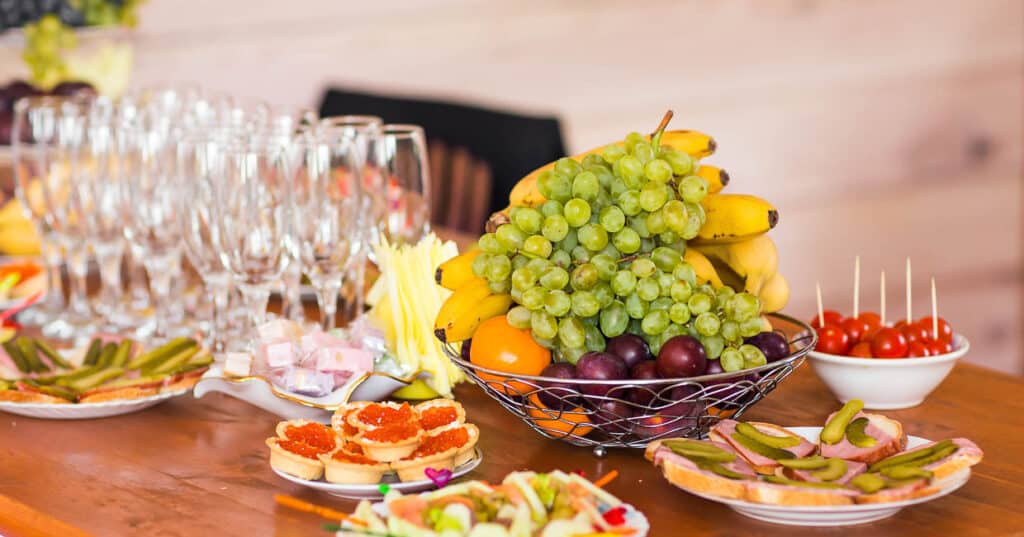 A potluck buffet table with a fruit basket, appetizers, and wine glasses arranged for a gathering.