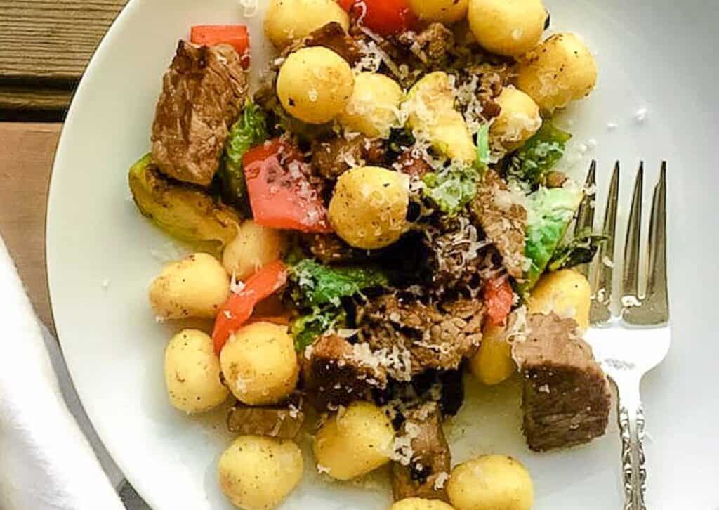 A plate of gnocchi with beef, vegetables, and grated cheese.