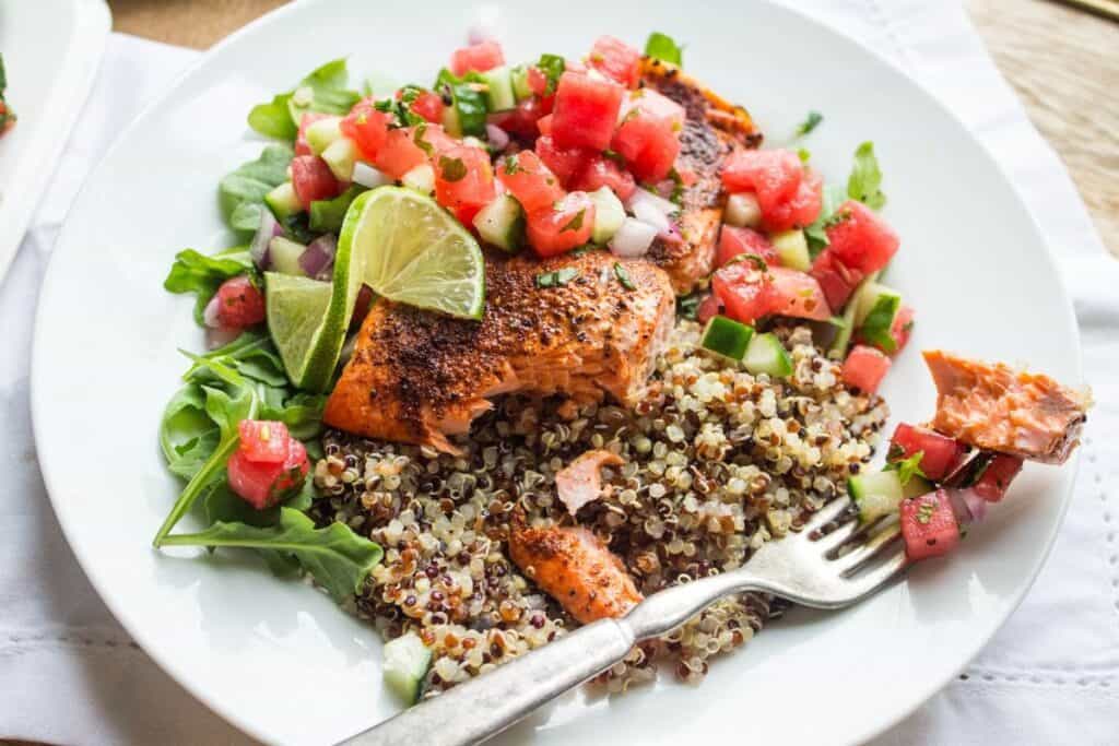 Grilled salmon fillet on a bed of quinoa with arugula, topped with fresh salsa and a lime wedge on a white plate.