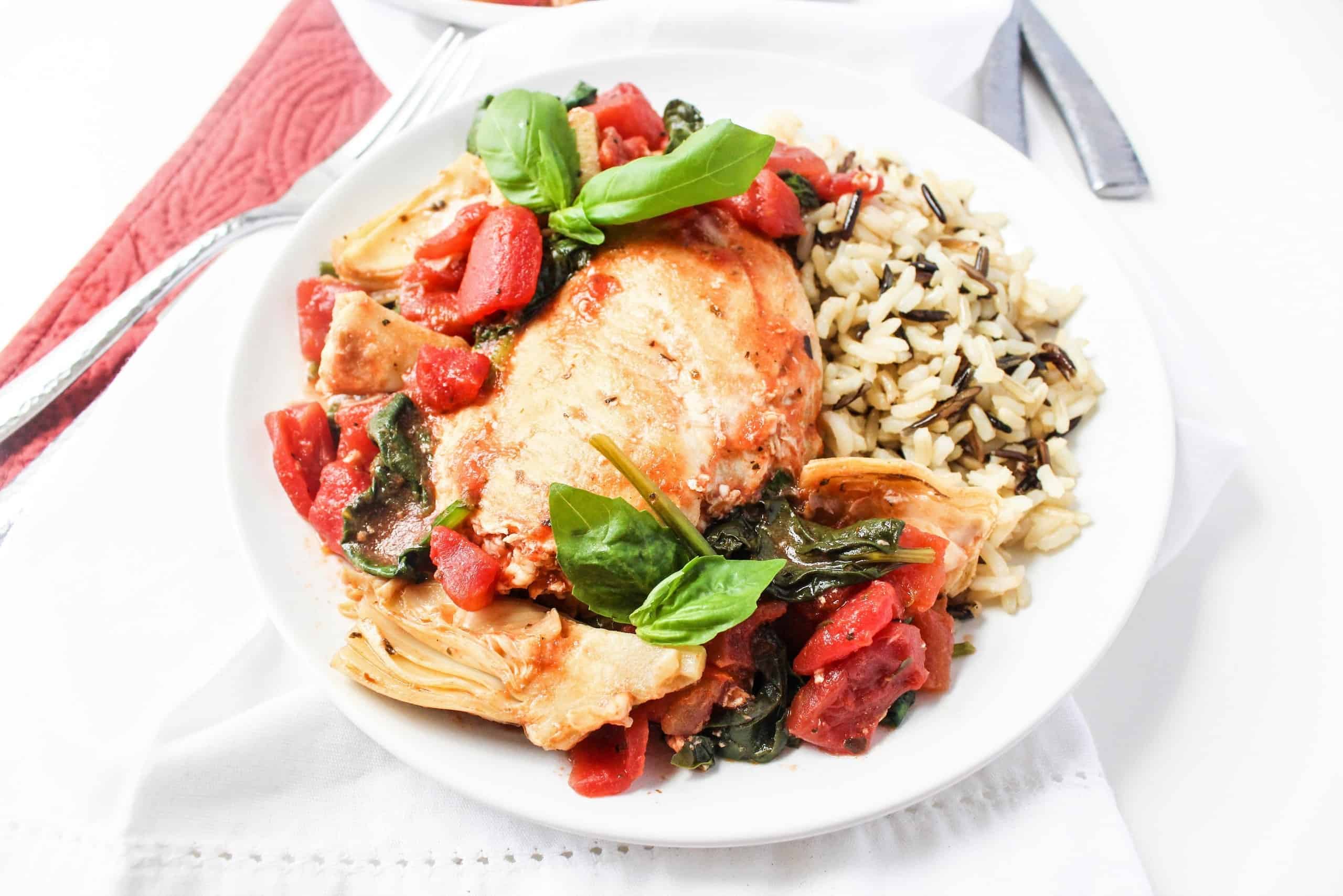 A white plate with chicken, stewed tomatoes, artichokes, sauteed spinach, and wild rice.