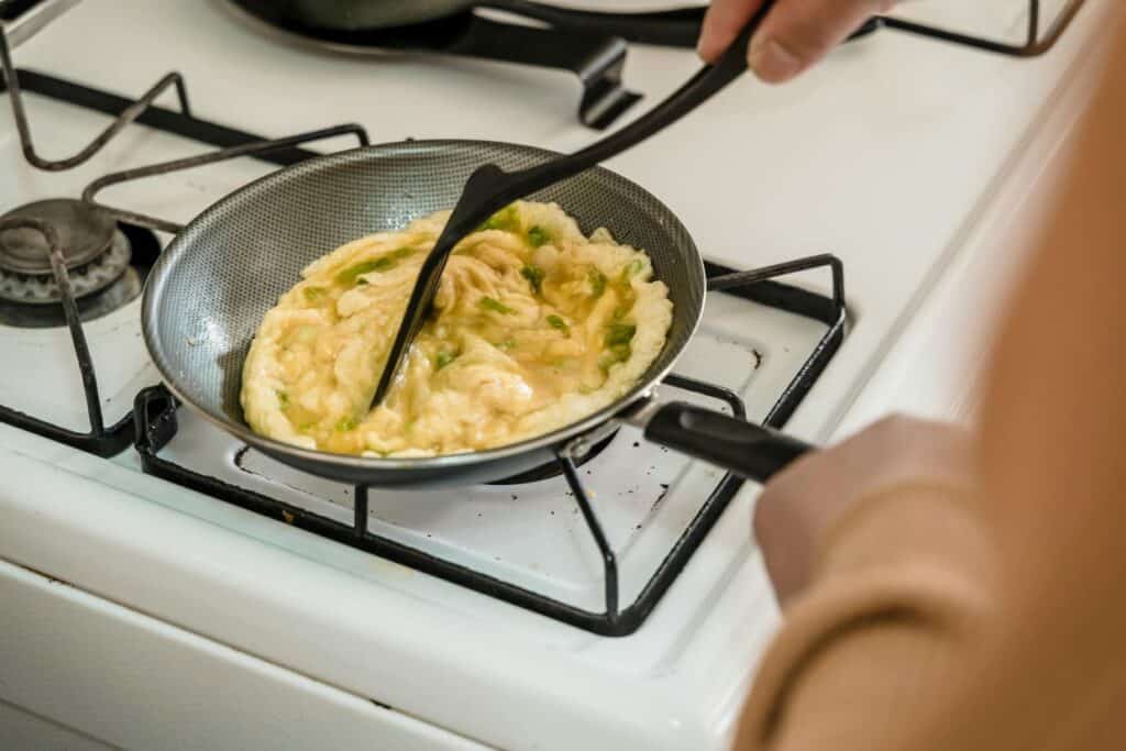 A person cooking an omelette in a frying pan on a stove, using a spatula to stir it.
