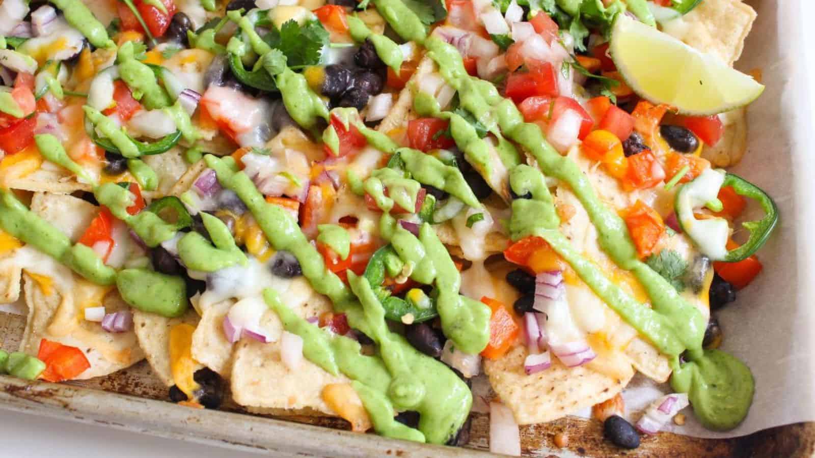 Cheesy nachos with black beans and diced bell peppers on a sheet pan with avocado crema drizzled on top.