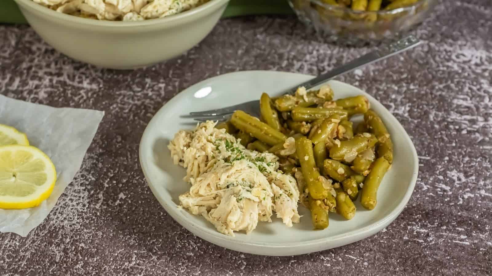 A plate of shredded chicken and green beans served with a garnish of herbs, accompanied by lemon slices and bowls of food in the background.