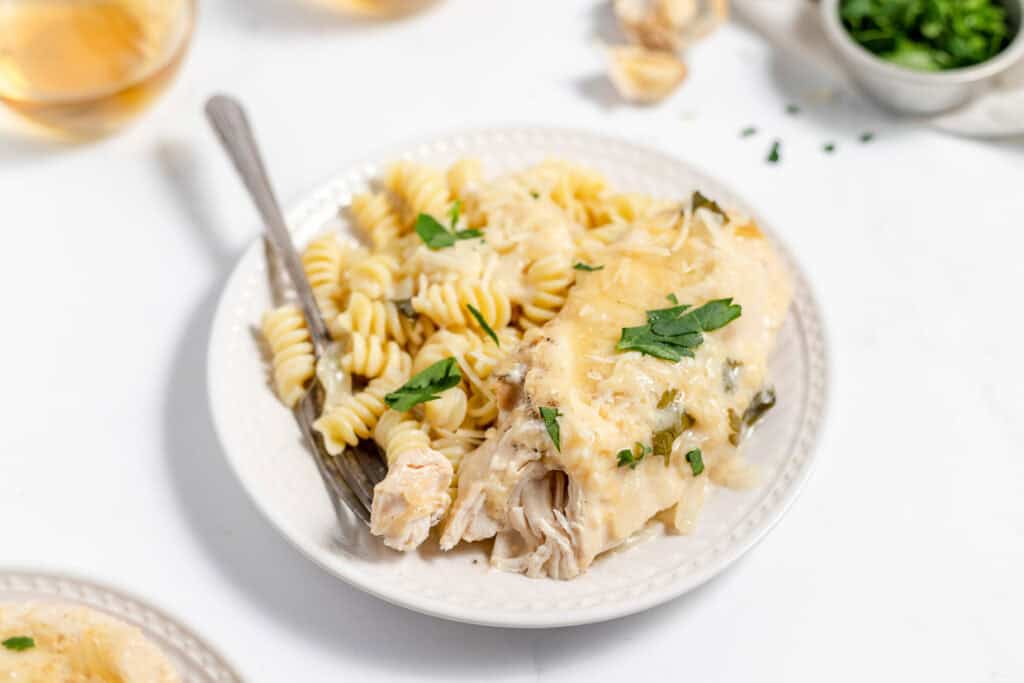 A plate of creamy chicken pasta topped with parmesan and herbs, with a fork on the side, on a white surface next to a bowl of dressing and greens.