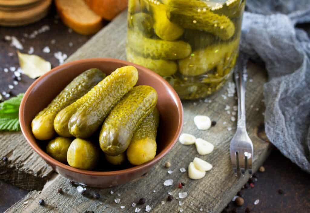A bowl of pickle flavor frenzy beside a jar and a fork, on a rustic wooden table with scattered salt and garlic cloves.