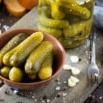 A bowl of pickle flavor frenzy beside a jar and a fork, on a rustic wooden table with scattered salt and garlic cloves.