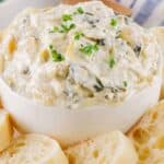 A bowl of creamy spinach dip garnished with chives, surrounded by sliced baguette pieces on a white and blue cloth.