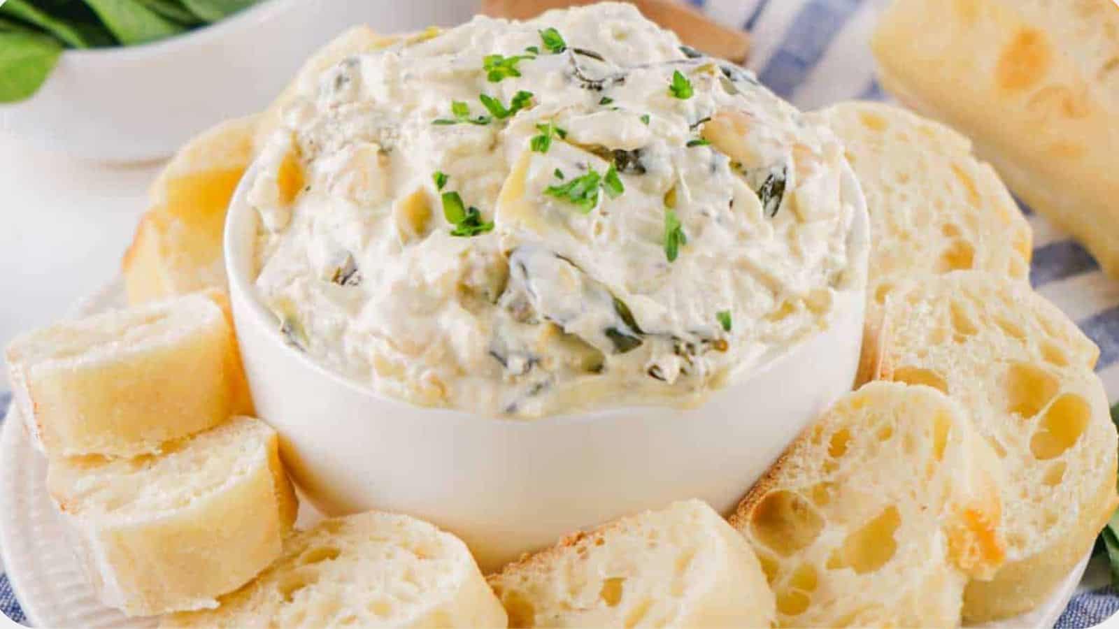 A bowl of creamy spinach dip garnished with herbs, surrounded by slices of crusty bread on a blue patterned cloth.