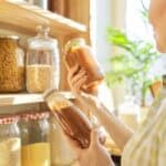 Woman in a pantry holding two jars of pasta sauce, with shelves stocked with various dry foods in clear containers.